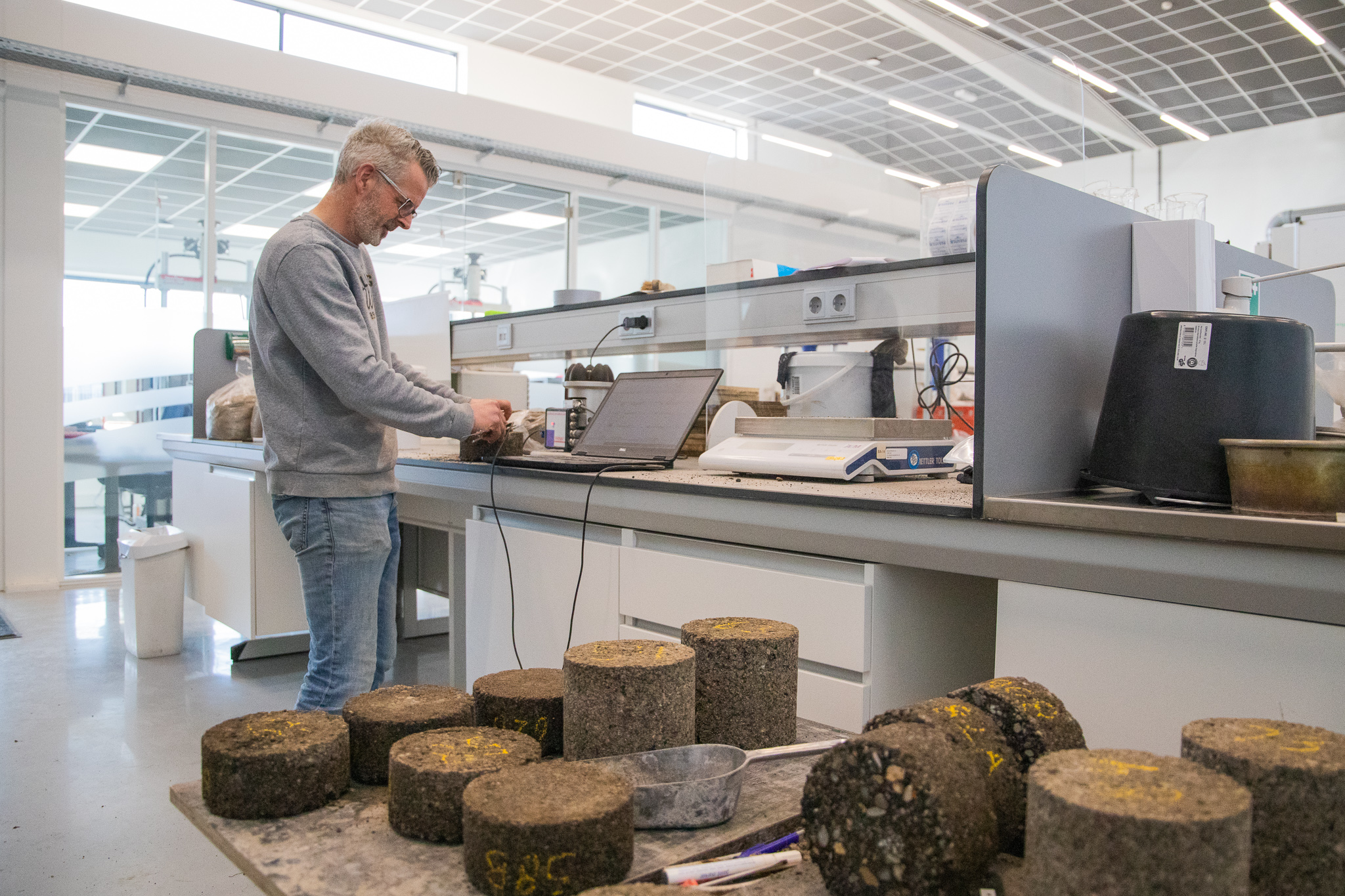 Man staat aan werkbank met laptop naast een aantal asfalt samples